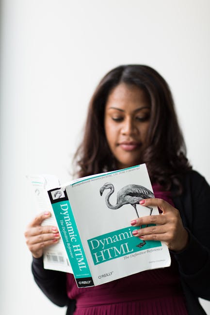 A woman with shoulder-length dark hair and a focused expression is holding an open magazine titled 'Dynamic HTML' published by O'Reilly. She is standing indoors against a plain white wall, dressed in