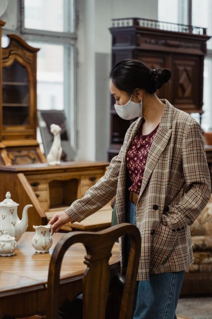 A woman wearing a face mask and a checked blazer is inside a room with large windows, lifting or inspecting a decorative porcelain teapot on a wooden dining table. The room features antique-style wood